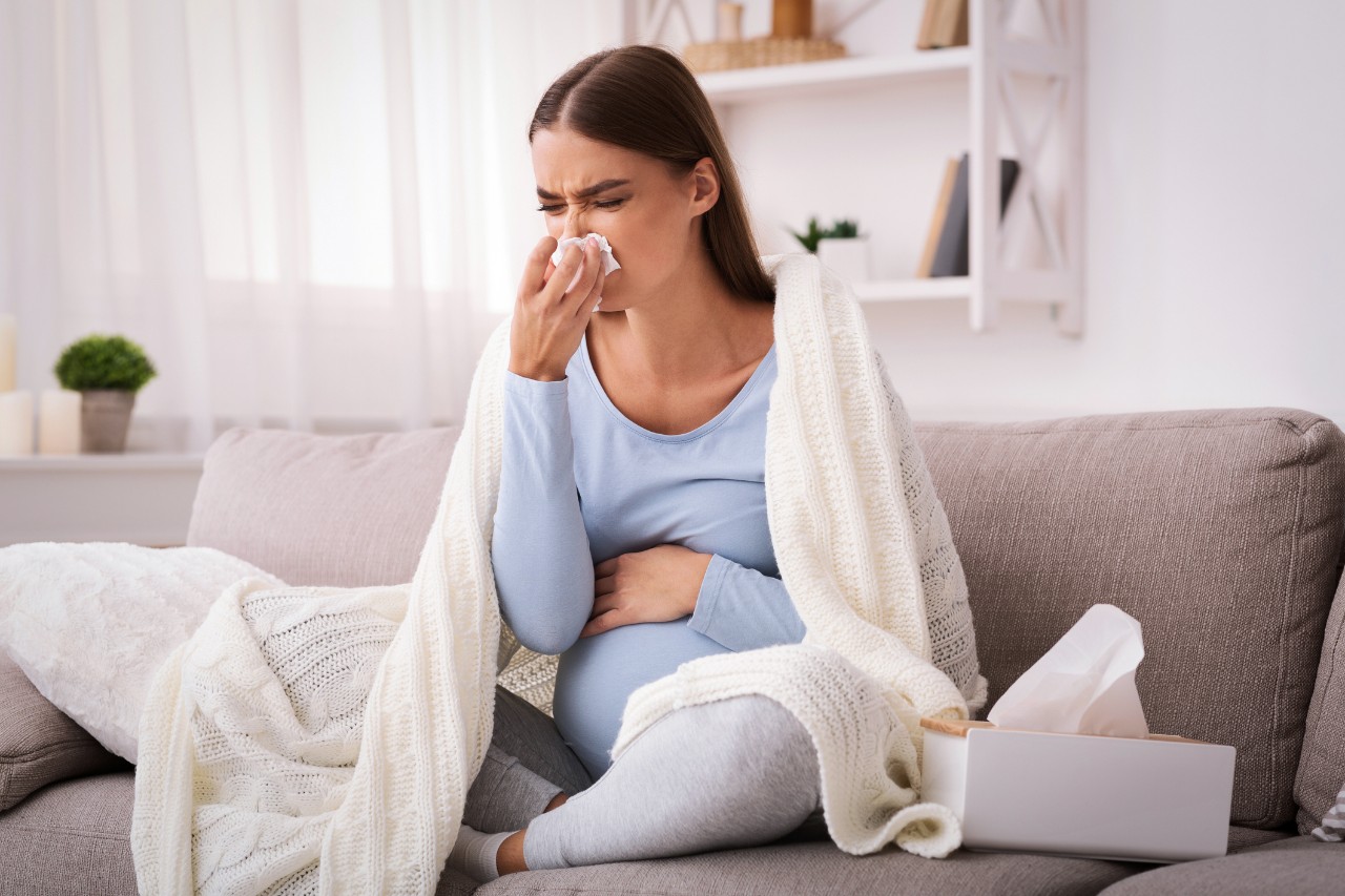 Pregnancy and illness. Sick pregnant woman blowing nose in tissue having fever sitting on sofa indoor