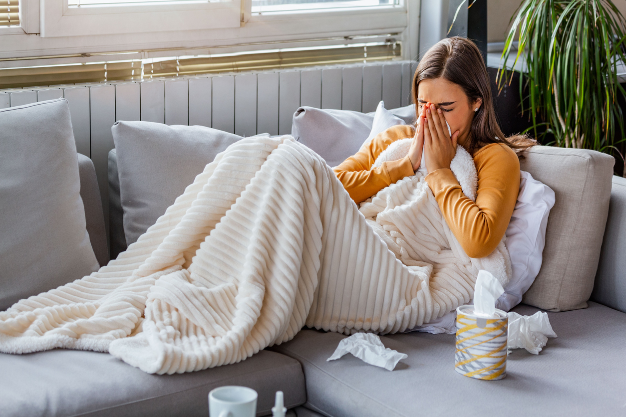 <p>Woman with a cold sneezing into a tissue, laying with a blanket on a couch</p>