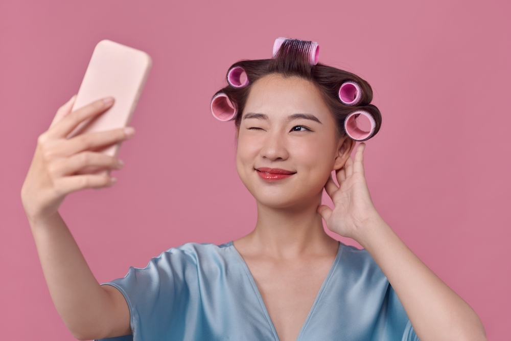 <p>Happy woman with hair curlers winking and taking a selfie with a pink background</p>