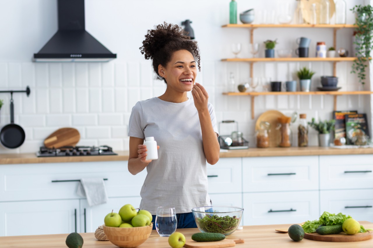 A woman stands at a kitchen island taking supplements and preparing healthy food.